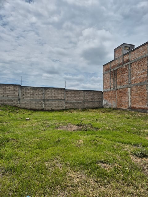 Abandoned concrete block building with brick tower structure in overgrown grassy field under cloudy sky